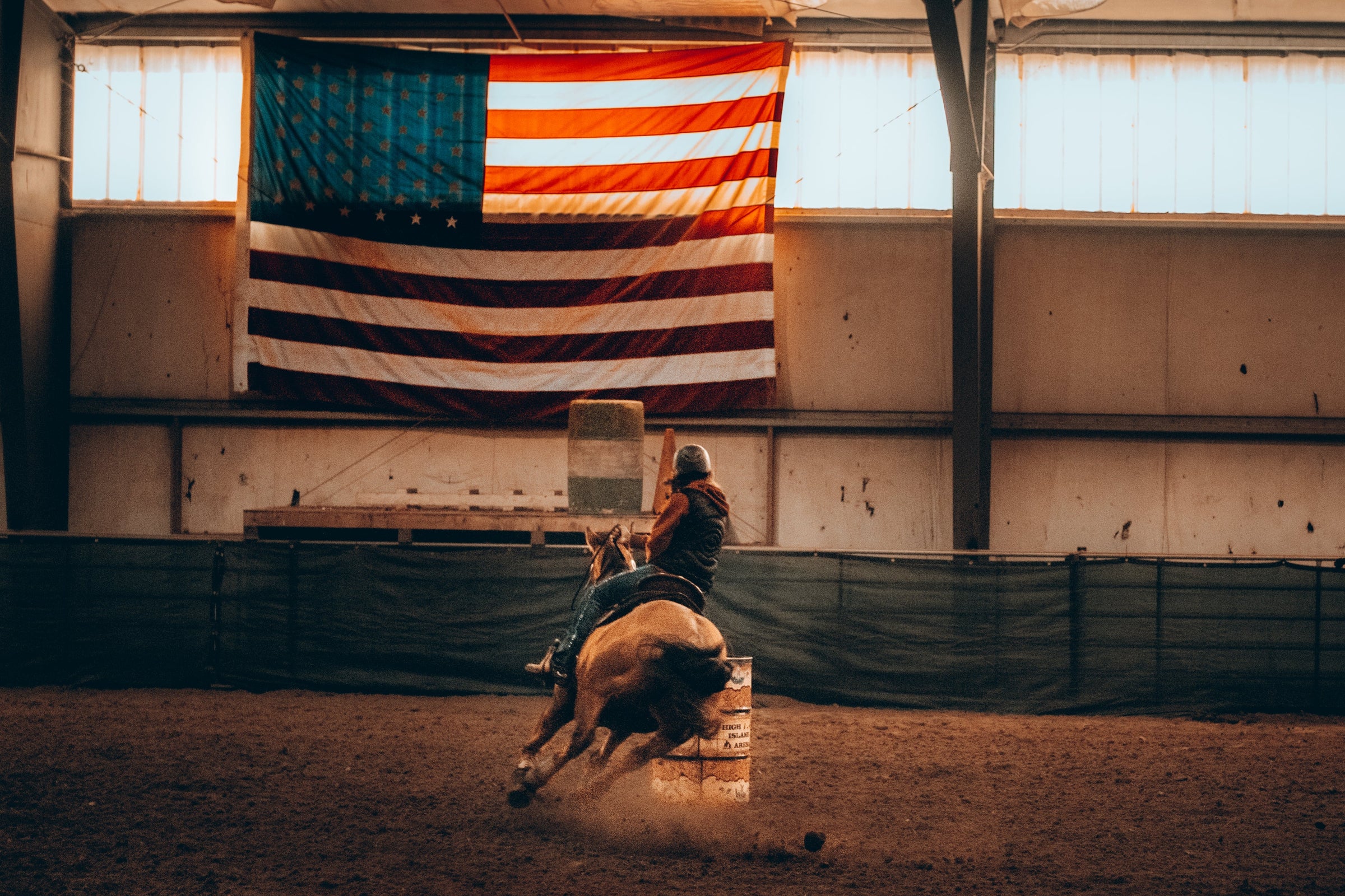 Person riding a bucking horse in an indoor arena with an American flag on the wall.