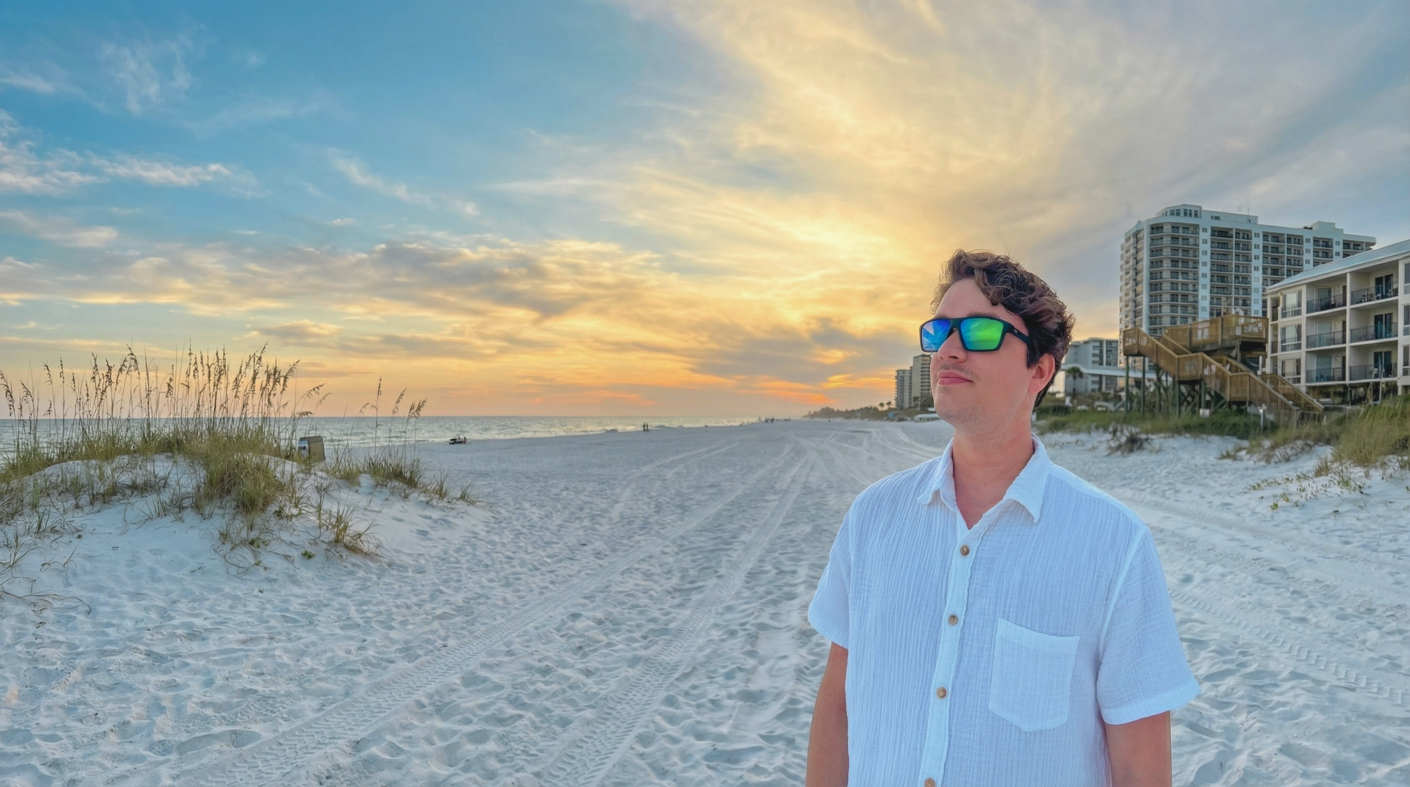Person wearing sunglasses on a beach at sunset with buildings in the background